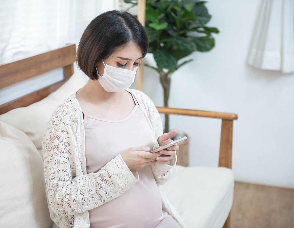 seated pregnant woman wearing mask looks at smart phone