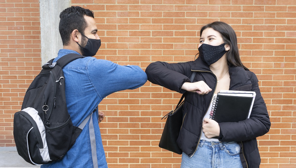 A man in his 20s and a woman in her 20s bump elbows in a greeting. They are both wearing black face masks. The man is standing on the left and is wearing a black backpack and denim collared shirt. He has black hair. The woman is standing on the right, has long black hair and is wearing a black coat and jeans. She is holding a black spiral notebook in her left arm.