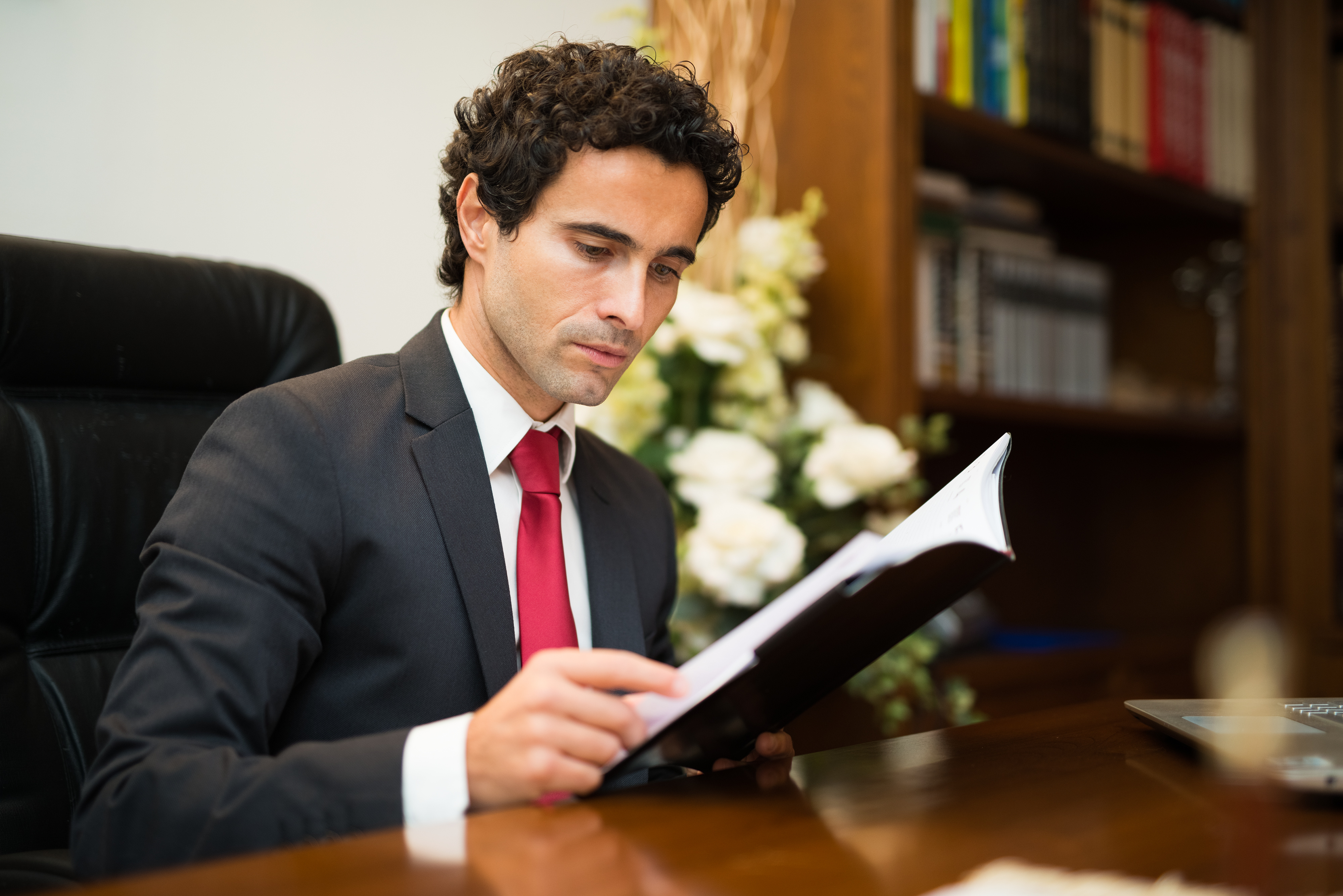 A business man with brown curly hair and light skin wearing a black suit, white shirt, and red tie sits at a desk reading a book. In the background are white flowers in a large vase and a bookshelf.