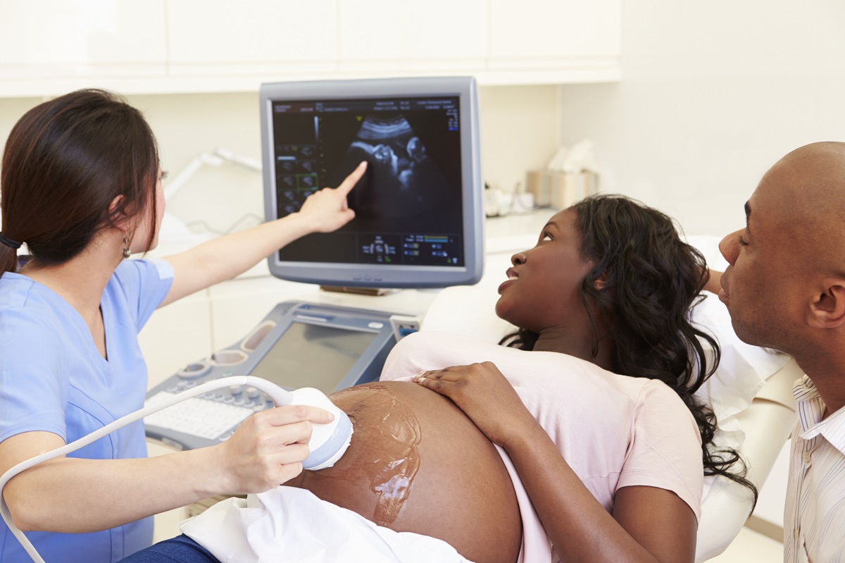 A pregnant woman with dark skin lays on a table while an ultrasound technician holds an ultrasound monitor to her stomach and points at a monitor. A man with dark skin sits next to the woman and observes.