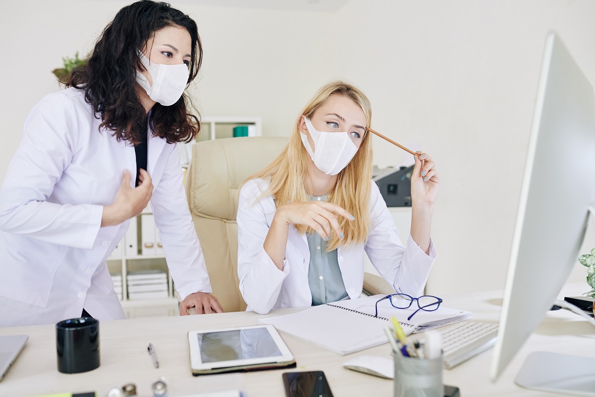 Two female doctors look at a computer screen on a desk. One is blonde and the other is brunette, both are wearing white lab coats and masks.