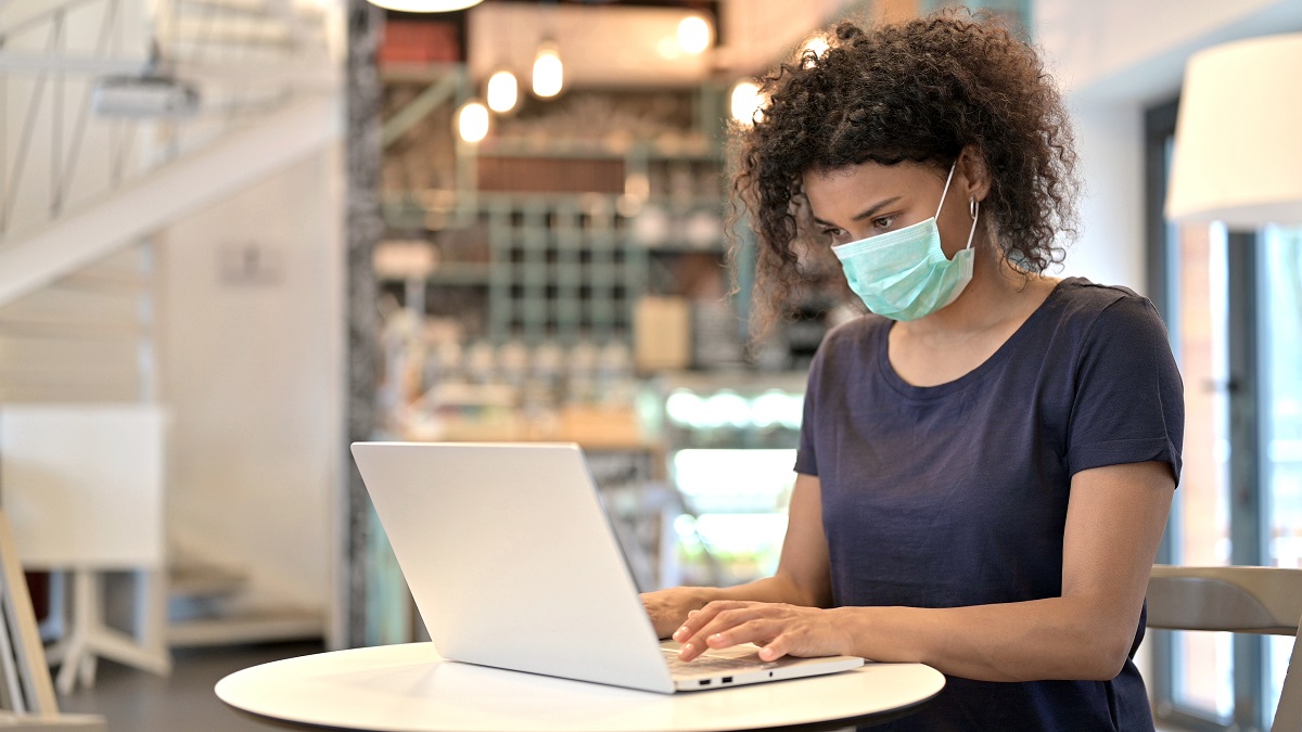 A woman with light brown skin wearing a medical mask sits at a table and looks at a laptop.