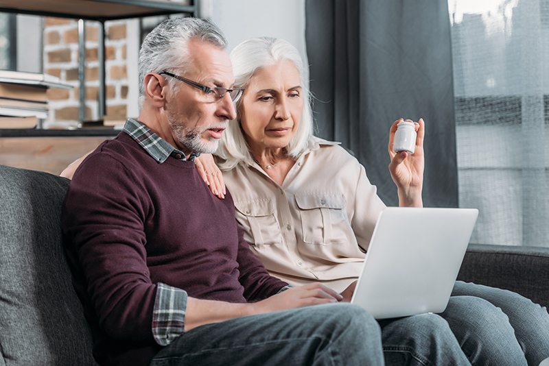 An older man and woman look at a laptop while sitting on a couch. The woman holds a pill bottle.