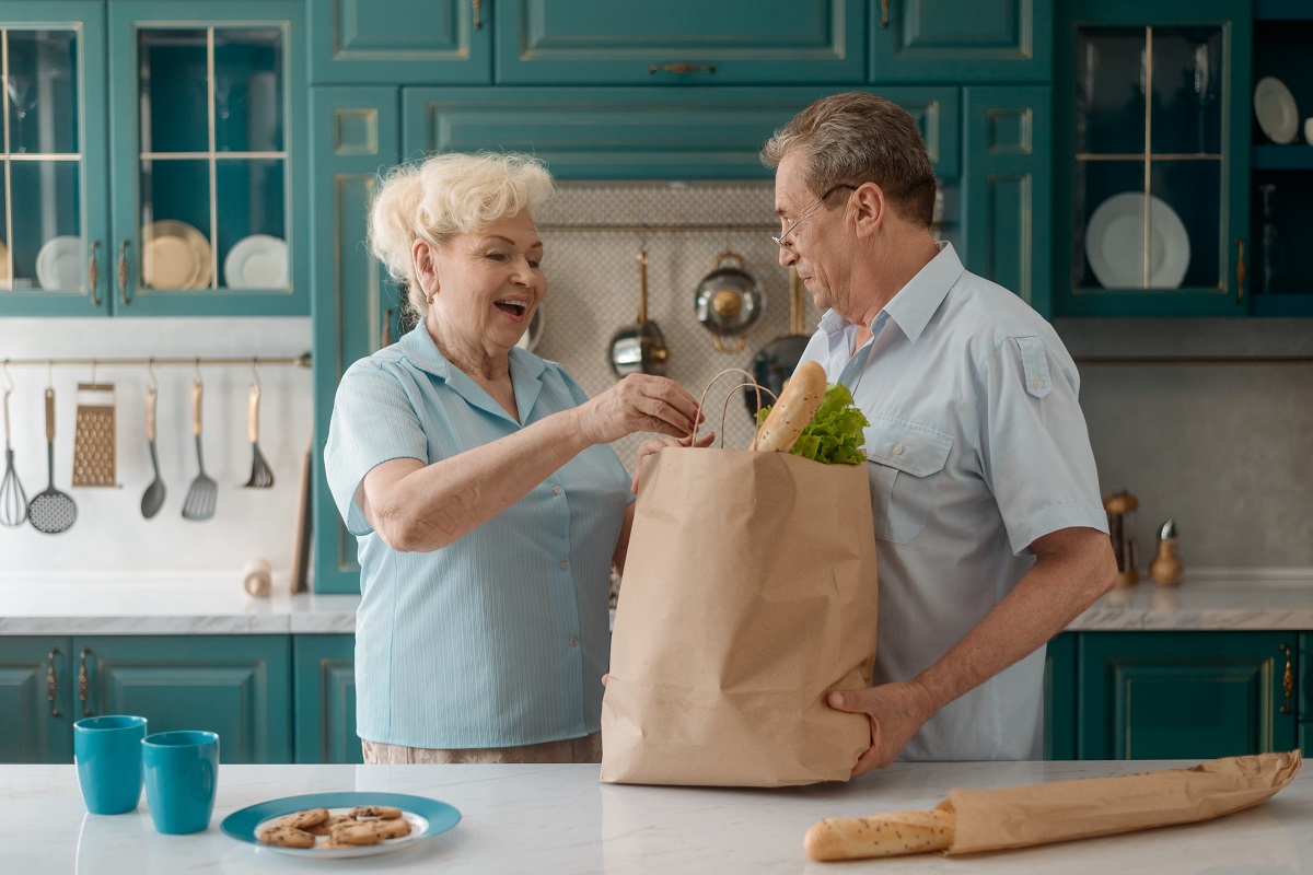 An elderly white couple smiles as they unpack groceries from a paper bag on their kitchen counter.
