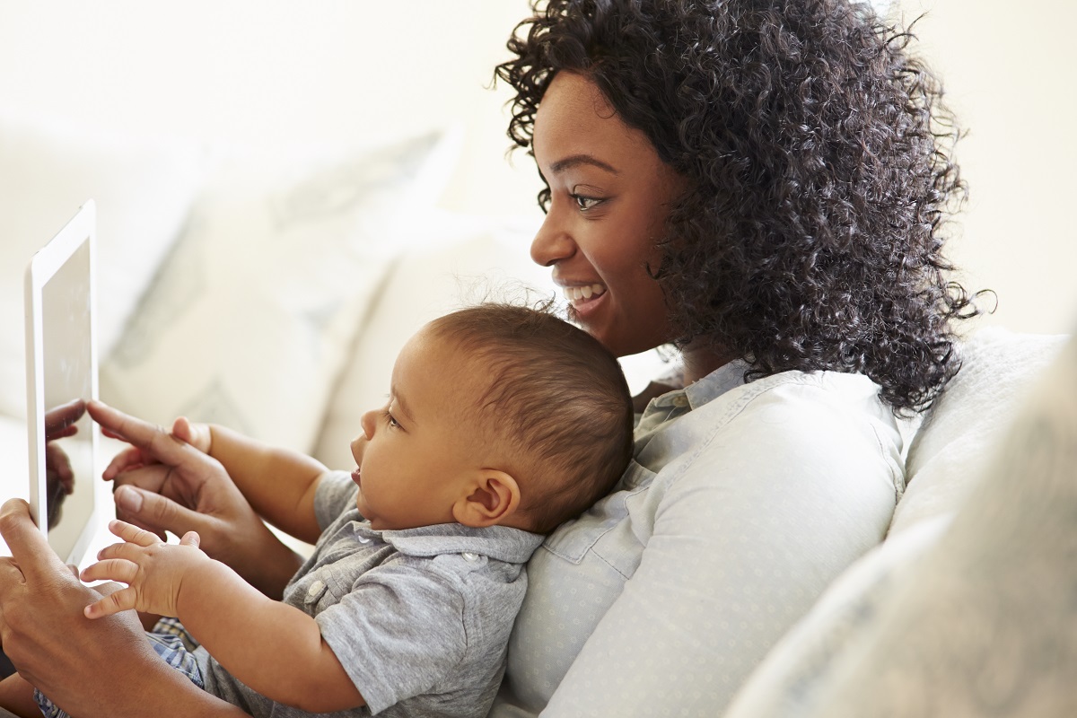 A woman with brown skin and curly hair holds a baby with brown skin. They both wear grey shirts and look at a tablet device while sitting on a couch.