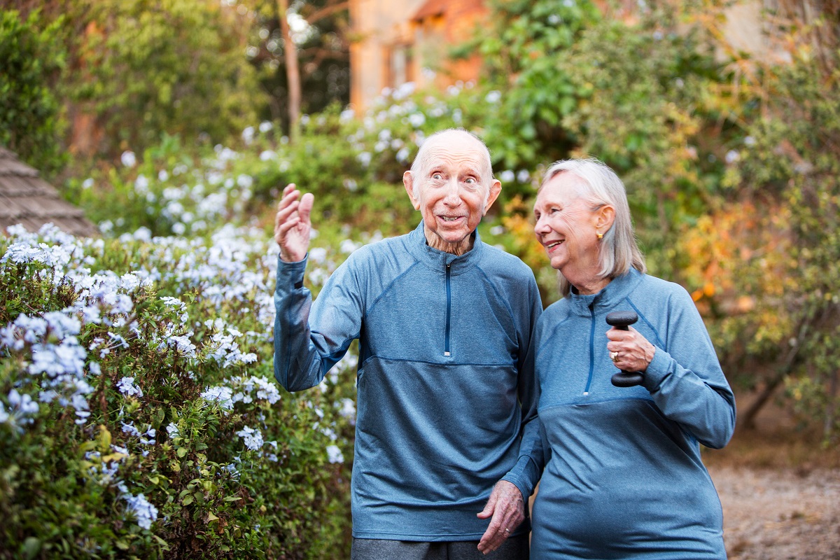 An older man and an older woman walk through a garden wearing matching blue sweaters