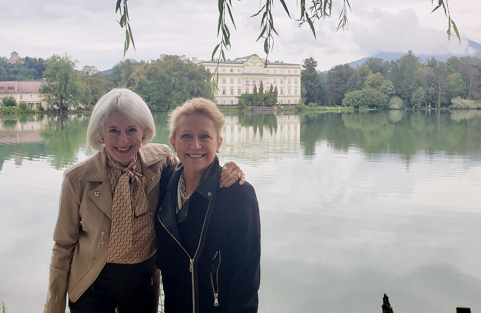 Karen Wolk Feinstein stands to the left of Lisa Simpson in Salzburg, Austria, 2019. Karen wears a tan moto jacket and tan top with tan scarf, and has short white hair. Lisa has long blonde hair tied back and wears a black moto jacket. In the background is a body of water and a white building on the opposite shore. The sky is cloudy.
