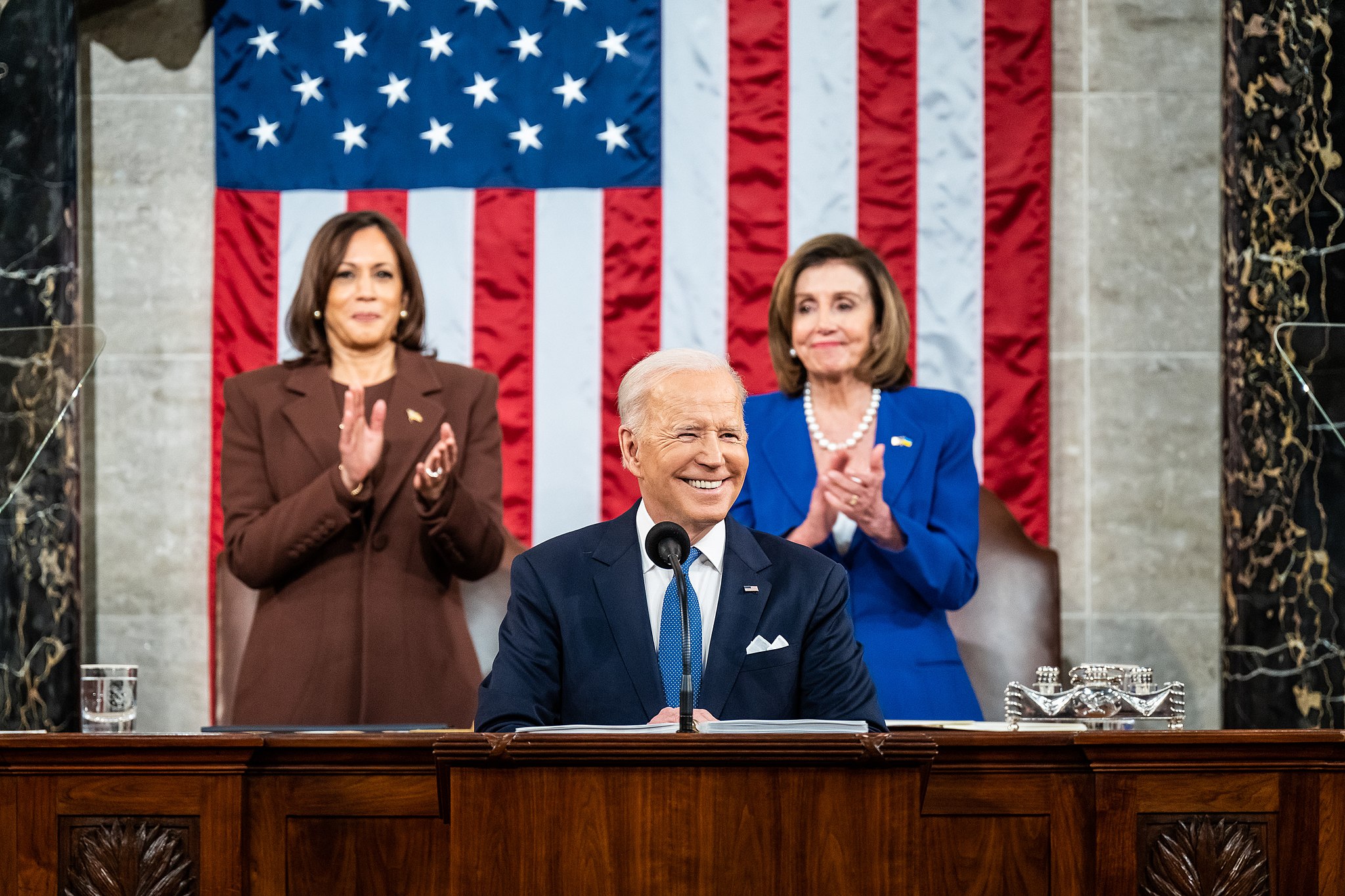 President Biden speaks at the state of the union address. Vice President Kamala Harris and Nancy Pelosi stand behind him while clapping. An American flag hangs on the wall behind them