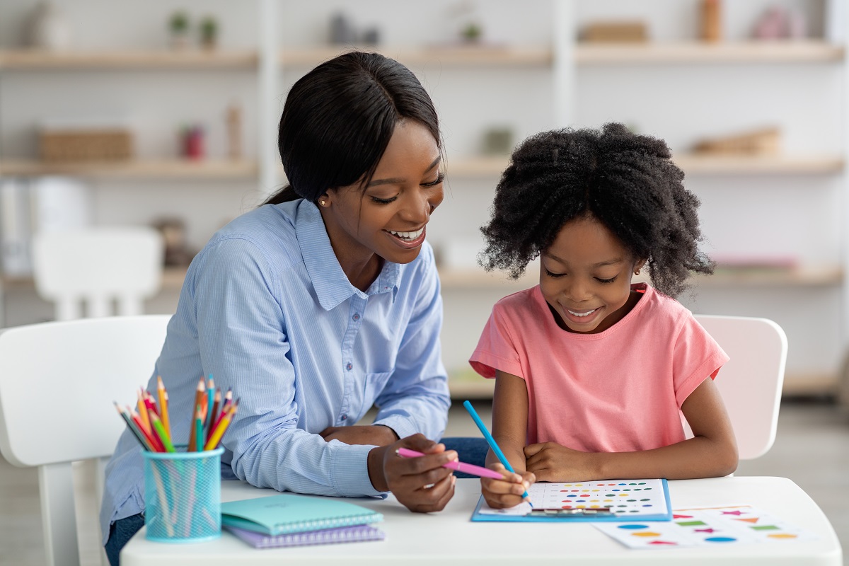 A counselor works with a young girl on an activity at a table. They both have dark brown skin. The counselor wears a blue button down shirt and the girl wears a pink tshirt.